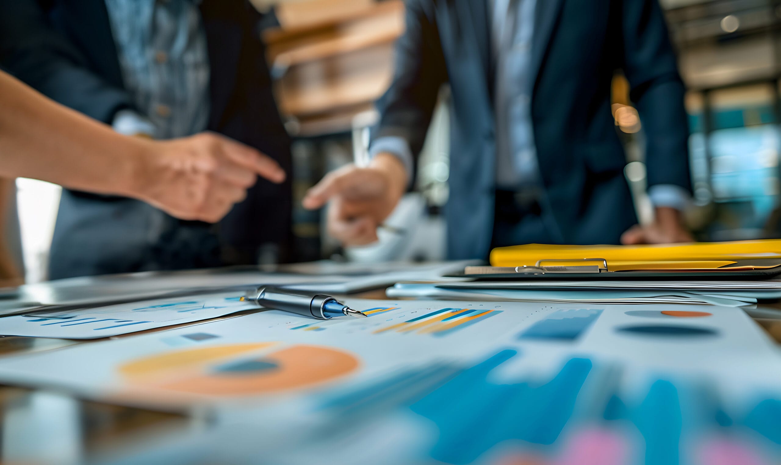Close-up of a business meeting with group of people in business attire pointing at a table covered with documents and colorful charts. A pen rests on top of the papers, emphasizing the focus on data and analysis. The blurred background captures the office setting with warm lighting, adding depth to the image and highlighting the collaborative atmosphere.
